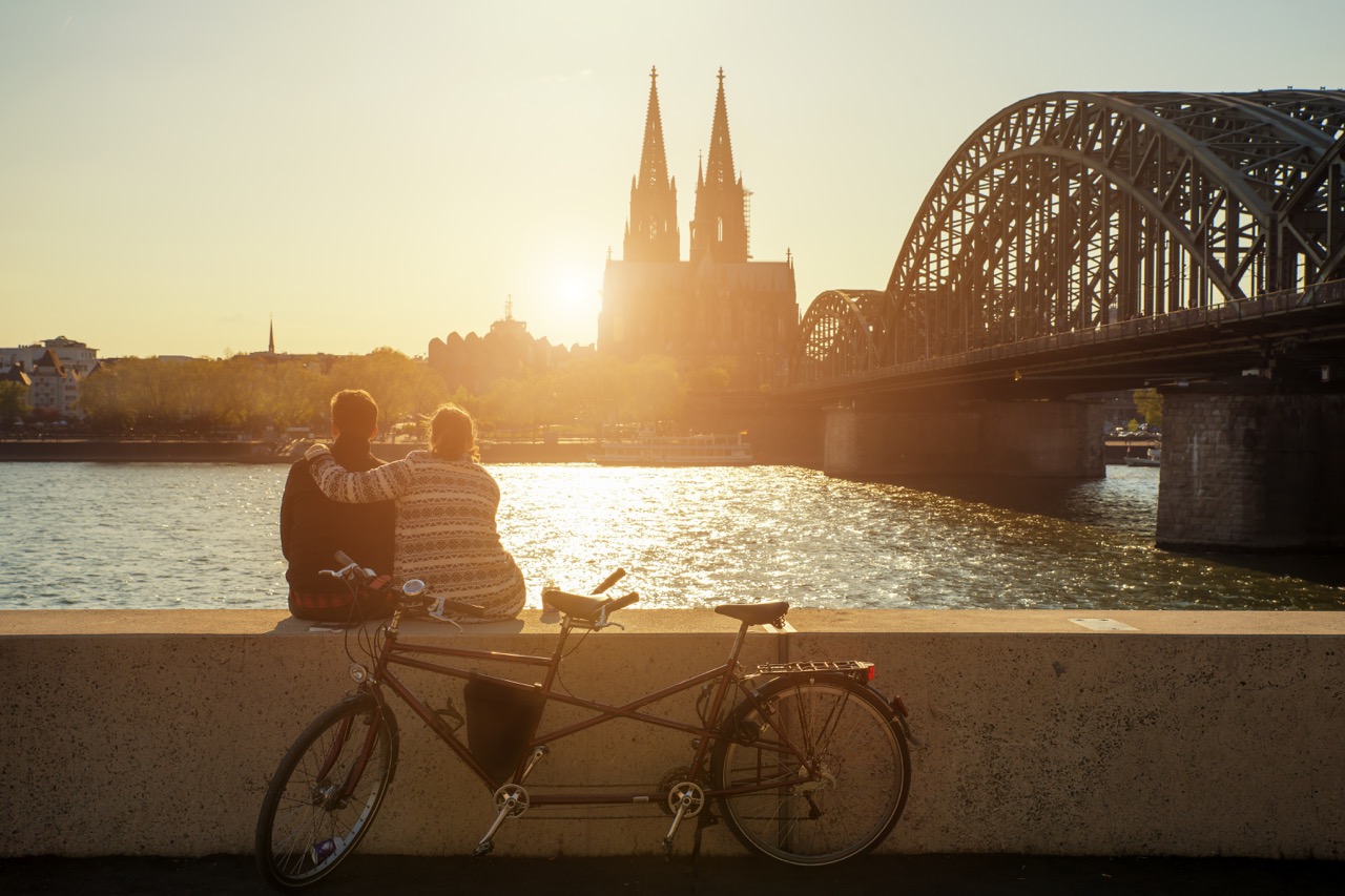  Ein Pärchen sitzt auf einer Mauer und schaut sich den Sonnenuntergang mit Blick auf den Kölner Dom an. 