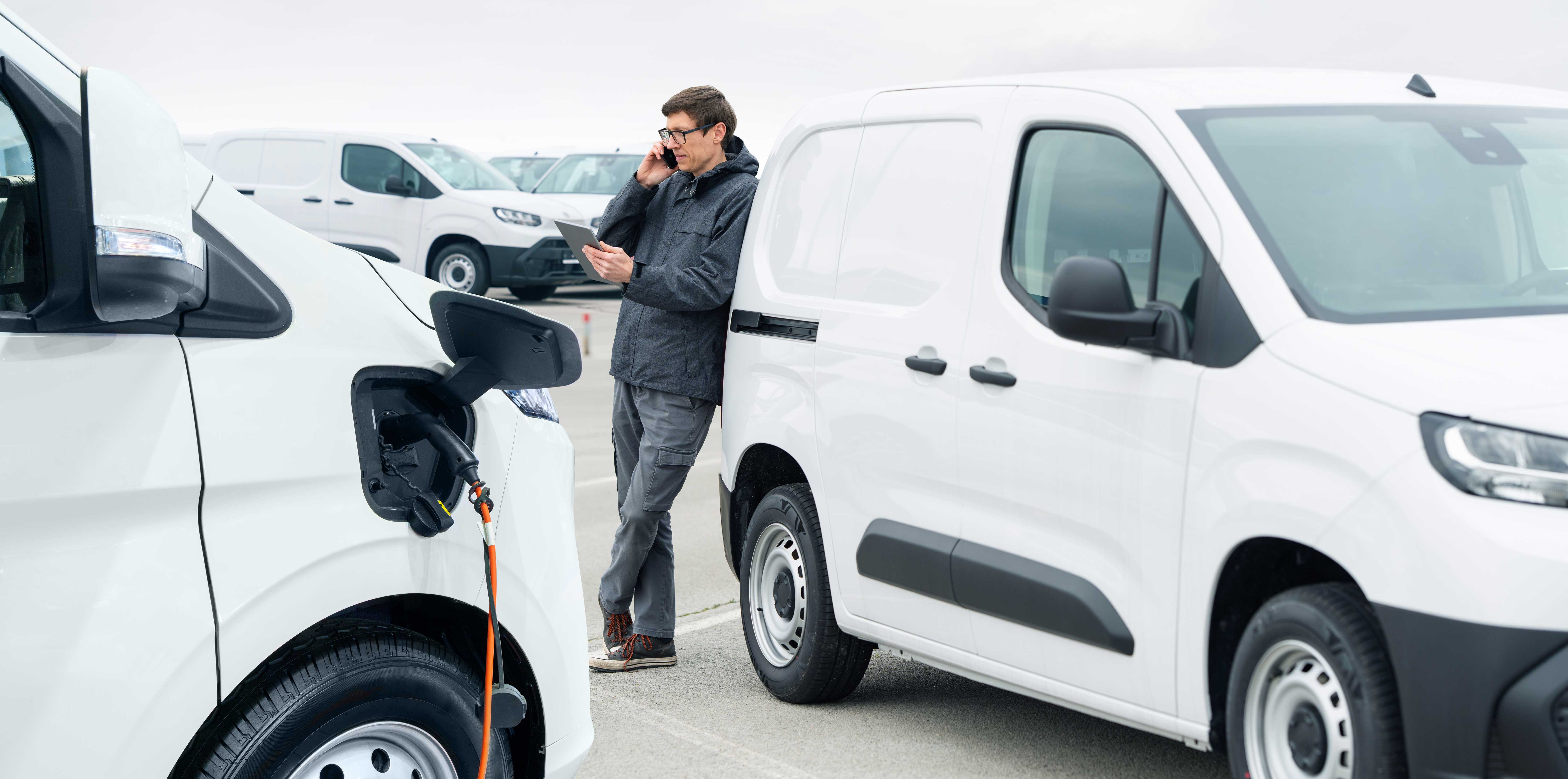  Mann telefoniert am Smartphone mit Tablet in der Hand an einen E-Transporter gelehnt. 