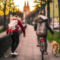 Im Sonnenuntergang fährt eine junge Frau auf dem Fahrrad mit der Leine von einem Hund in der Hand. Neben ihr fährt eine weitere junge Frau auf dem Skateboard.