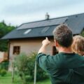 Vater mit Kleinkind auf dem Arm zeigt auf ein Einfamilienhaus im Grünen.