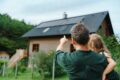 Vater mit Kleinkind auf dem Arm zeigt auf ein Einfamilienhaus im Grünen.