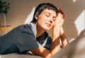  Junge Frau liegt auf dem Bett und stützt ihren Kopf seitlich mit den Händen. Die Sonne scheint ihr ins Gesicht. Sie hat die Augen geschlossen und lächelt. 