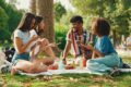 Freundesgruppe, die auf einer Picknickdecke im Park sitzt und gemeinsam isst und spricht.