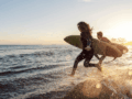 Zwei Menschen rennen mit ihrem Surfbrett bei untergehender Sonne ins Meer.