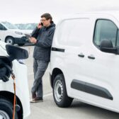 Mann telefoniert am Smartphone mit Tablet in der Hand an einen E-Transporter gelehnt.