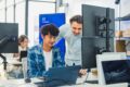 Ein Team für die gemeinsame Entwicklung von KI in einem modernen Tech-Büro: Ein junger Entwickler programmiert an seinem Laptop, während sich ein erfahrener Kollege über ihn beugt und mit ihm über Code und Datenvisualisierungen auf mehreren Monitoren spricht.
