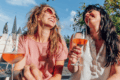 zwei Frauen sitzen draußen in der Sonne, trinken Wein und lachen