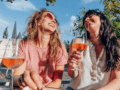 zwei Frauen sitzen draußen in der Sonne, trinken Wein und lachen